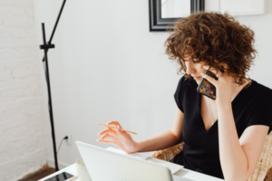 Image of a woman on a mobile phone. The woman is sat at a desk in front of a laptop. She is holding a phone in her left hand and a pen in her right hand.
