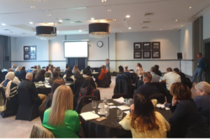 People in a room sat round tables listening to a presentation at a Proactive Care event
